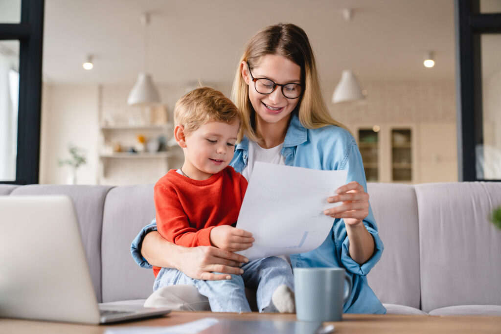 Little small toddler kid child son helping her busy young mother with freelance remote work holding papers, documents and domestic bills at home on lockdown during maternity leave