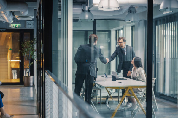 Business Leaders Complete a Job Interview With a Confident Handshake in a Glass Meeting Room, While a Hiring Manager Reviews Notes and Additional Candidates Wait in the Hallway